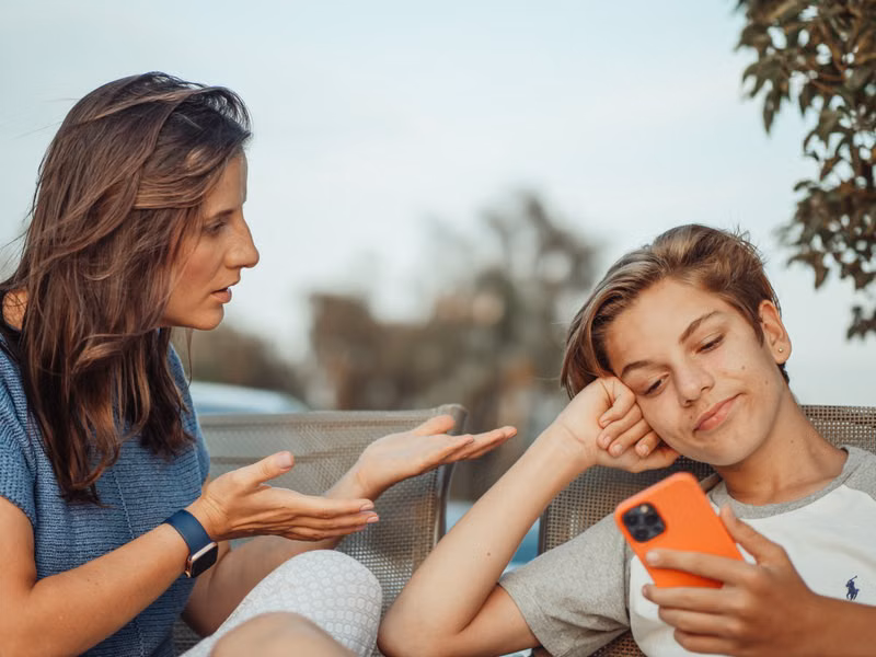 A mother engaging with her teenage son holding a smartphone outdoors.