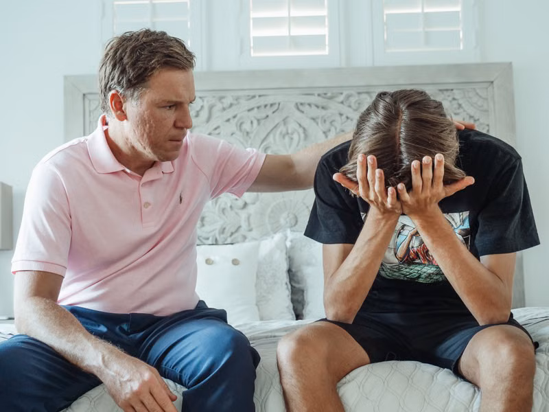 A compassionate father consoles his upset teenage son on a bed indoors.